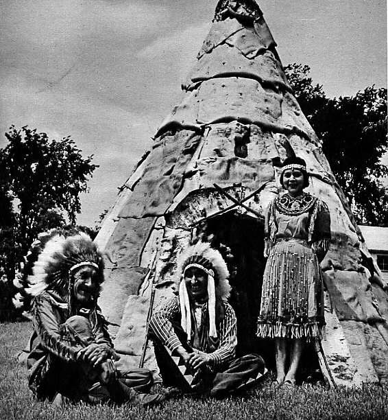 Trois Abénakis en costume traditionnel, devant leur wigwam